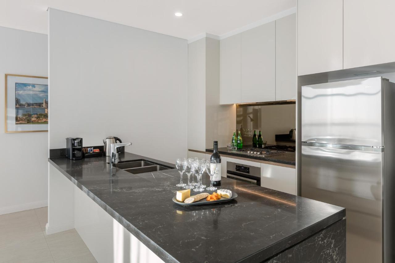 Meriton Suites Sussex Street, Sydney Exterior foto The photo shows a modern kitchen featuring a sleek design with a dark stone countertop. There is a built-in sink and possibly a cooktop. The kitchen cabinetry is light-colored, creating a contrast with the dark counter. On the countertop, there are w
